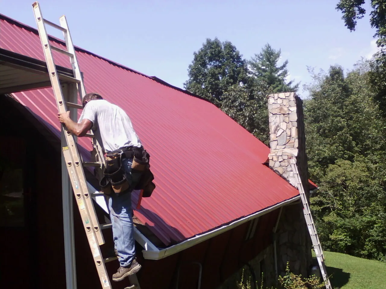 Roofer on a ladder beside a red metal roof and stone chimney — Soffit & Fascia Repair by Roofing Brothers in Fallston